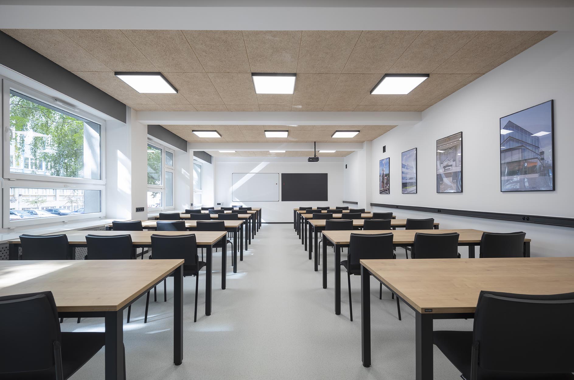 Pusta nowoczesna sala lekcyjna z rzędami drewnianych stolików, czarnymi krzesłami, dużymi oknami i tablicą