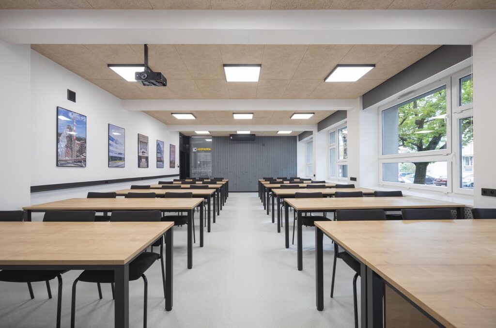 Pusta nowoczesna sala wykładowa z rzędami drewnianych stołów, czarnymi krzesłami, projektorem sufitowym i dużymi oknami