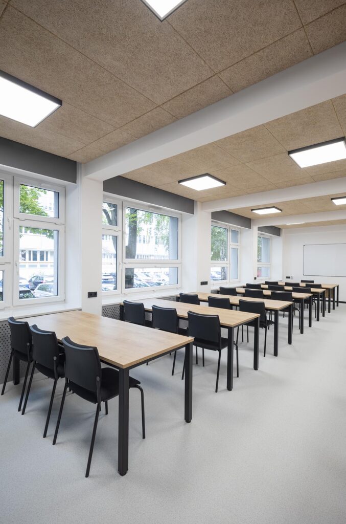 Pusta nowoczesna sala lekcyjna z rzędami drewnianych stołów i czarnych krzeseł, dużymi oknami i białą tablicą