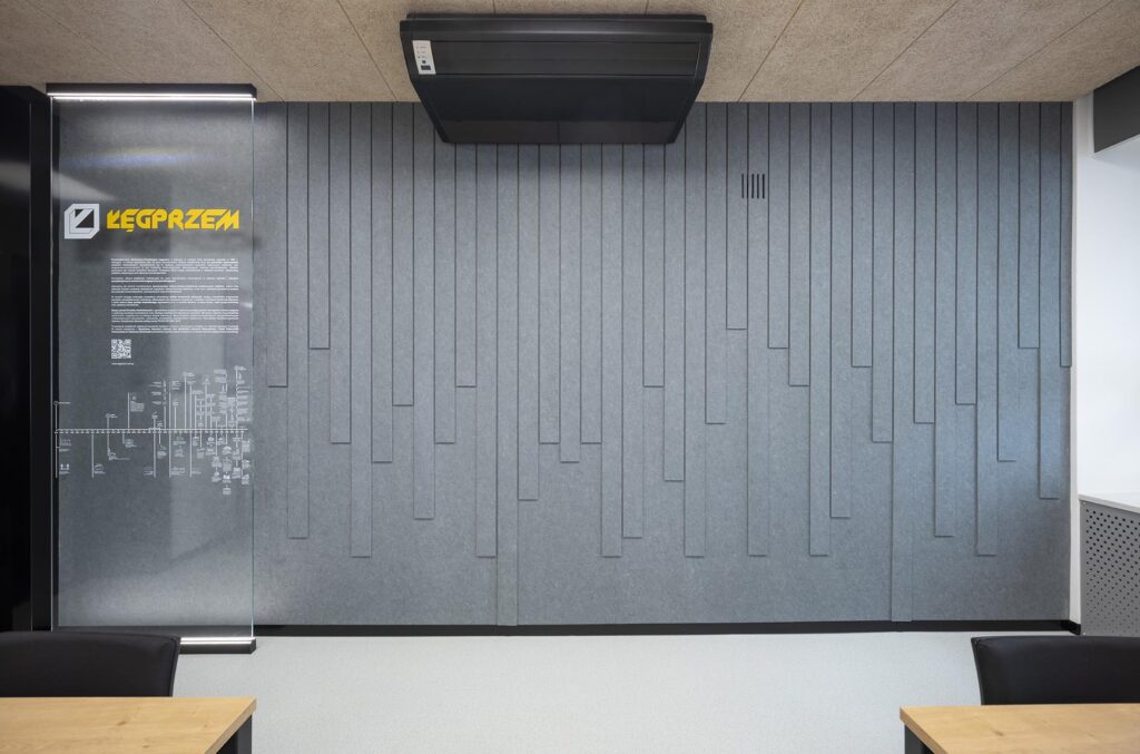 Nowoczesne wnętrze biurowe z szarą ścianą akustyczną o pionowych panelach, klimatyzatorem i szklaną tablicą z żółtym napisem, fragmenty biurek in situ