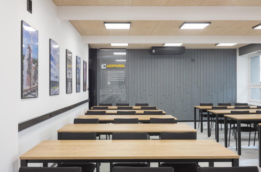 Pusta nowoczesna sala wykładowa z rzędami drewnianych stołów i czarnych krzeseł, obrazami na ścianie i panelami akustycznymi
