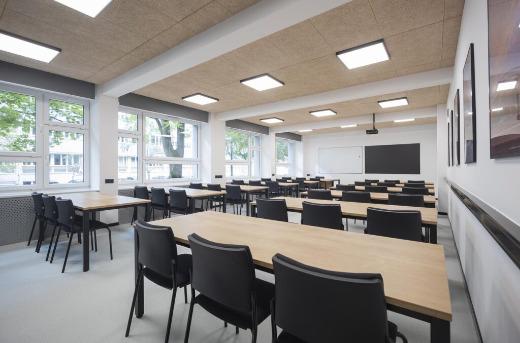 Pusta nowoczesna sala lekcyjna z rzędami drewnianych stołów i czarnych krzeseł, dużymi oknami, tablicą i projektorem