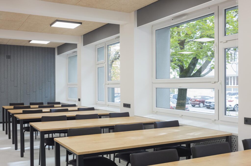 Pusta nowoczesna sala lekcyjna z rzędami drewnianych stołów i czarnych krzeseł oraz dużymi oknami z widokiem na drzewo i samochody