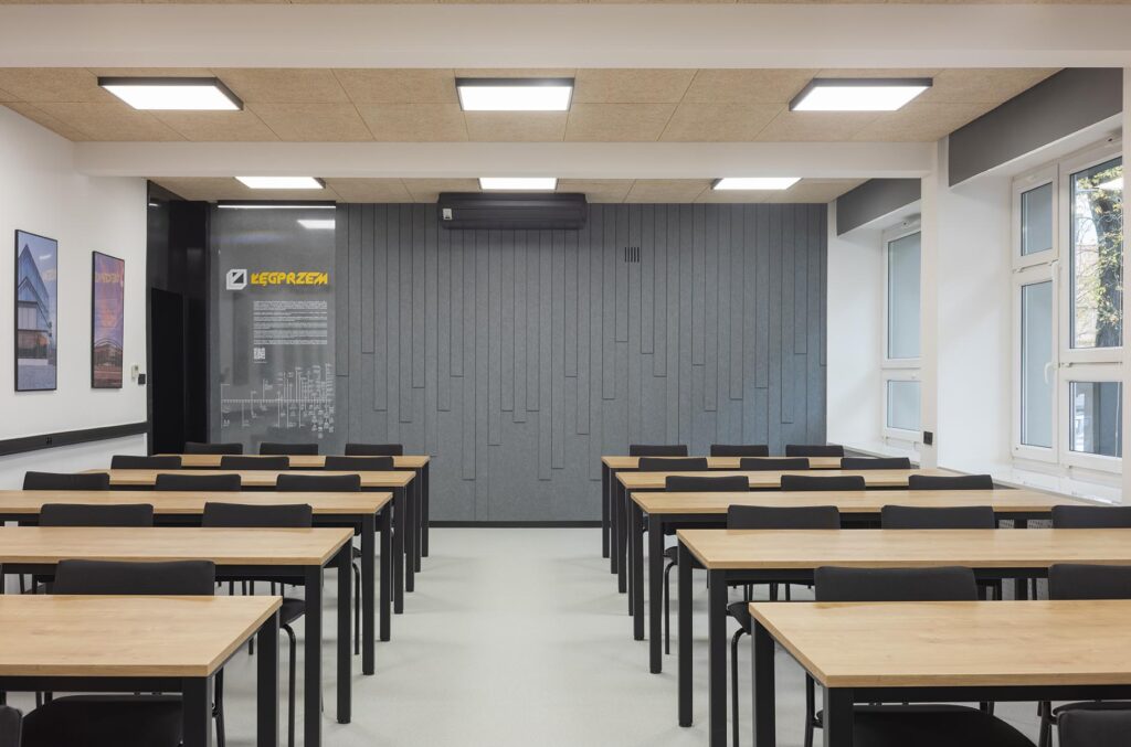 Pusta nowoczesna sala lekcyjna z rzędami drewnianych stolików i czarnych krzeseł, szarą ścianą akustyczną i dużymi oknami