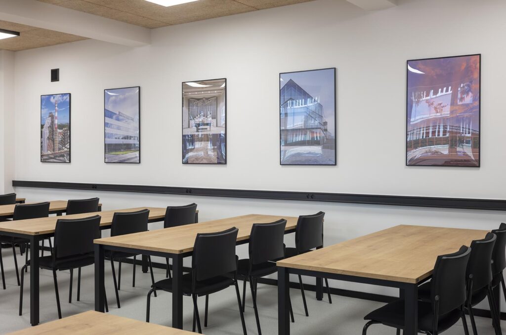 Pusta nowoczesna sala do nauki z drewnianymi stołami, czarnymi krzesłami i obrazami na białej ścianie