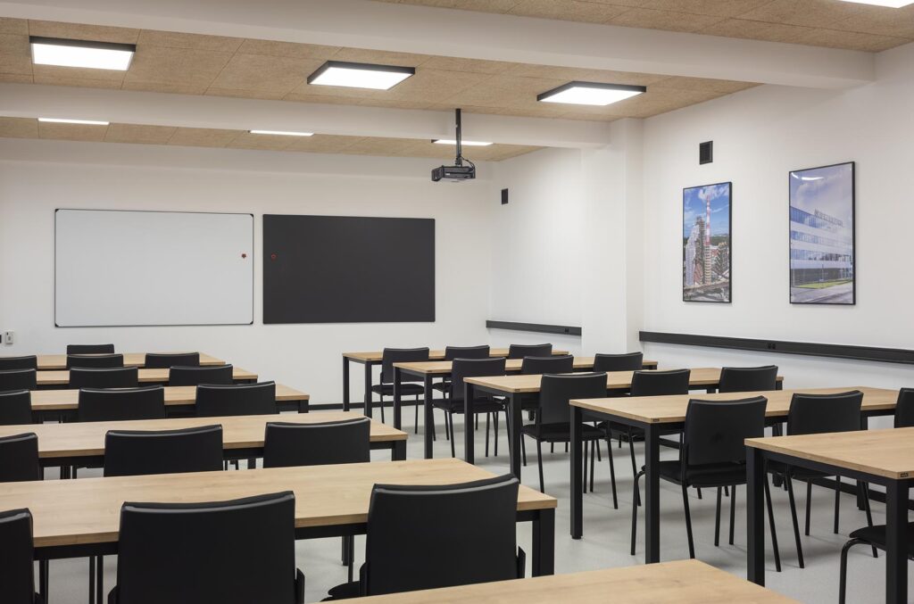 Pusta nowoczesna sala lekcyjna z rzędami drewnianych biurek, czarnymi krzesłami, tablicą i projektorem sufitowym