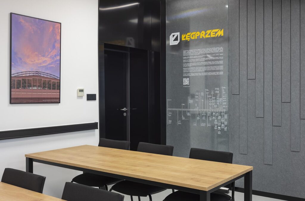 Sala konferencyjna z drewnianym stołem, czarnymi krzesłami, obrazem na ścianie i szarą przeszkloną ścianką z logo