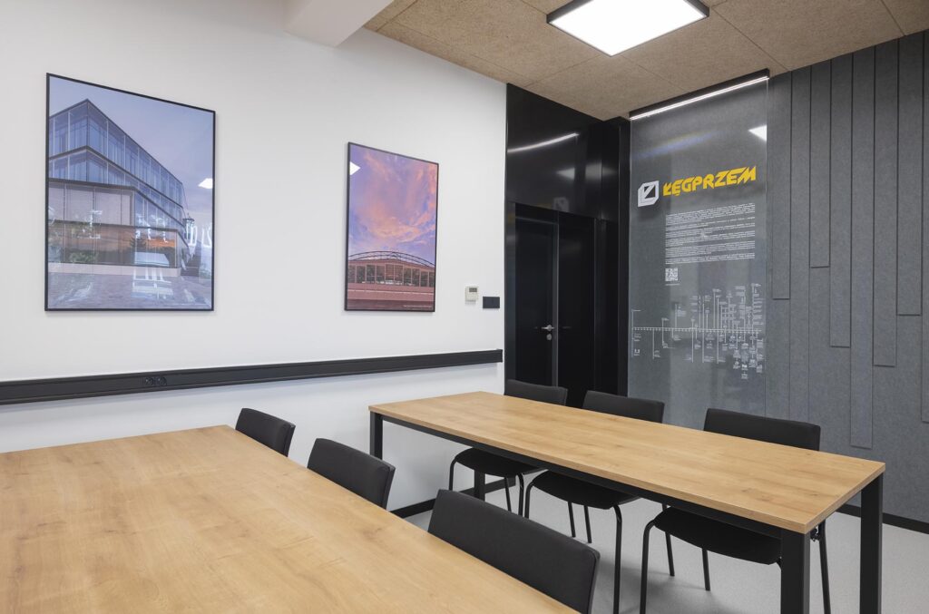 Mała sala konferencyjna z drewnianymi stołami, czarnymi krzesłami i plakatami na ścianie