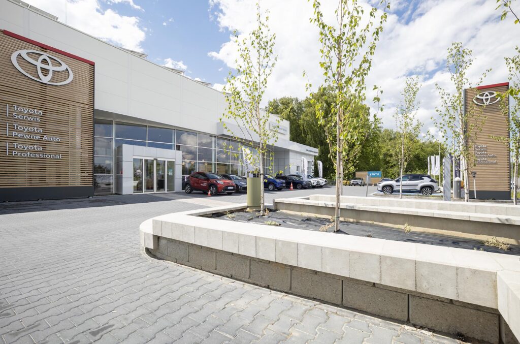 Salon samochodowy Toyota z przeszkloną elewacją, zaparkowanymi samochodami i klombami z młodymi drzewkami