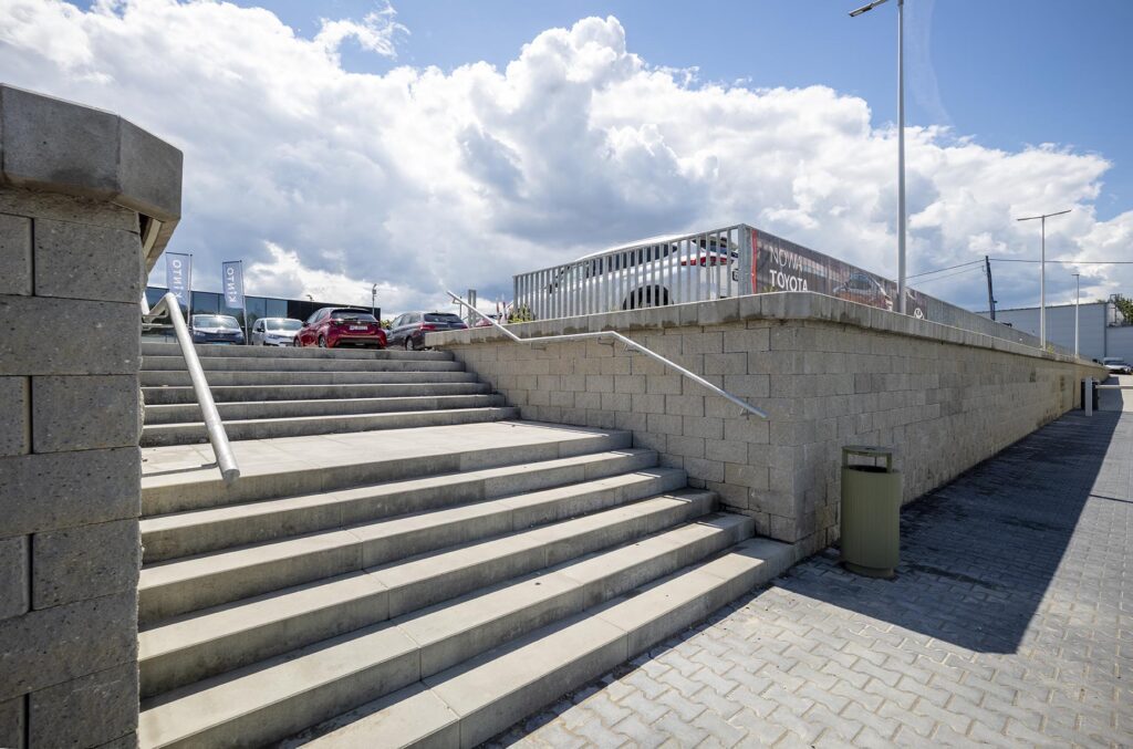 Betonowe schody z metalowymi poręczami prowadzące na parking z zaparkowanymi samochodami pod chmurzystym niebem