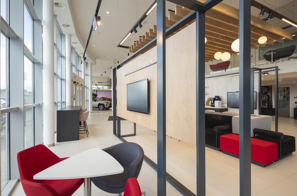 Nowoczesne wnętrze salonu samochodowego z dużymi oknami, strefą siedzącą z czerwonymi i ciemnymi fotelami oraz ekranem na drewnianej ściance