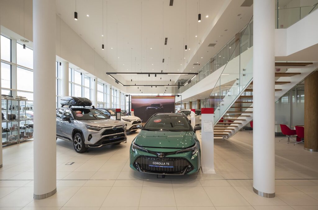 Salon samochodowy z zieloną Toyotą Corolla TS na środku, srebrnym SUV-em z bagażnikiem dachowym, schodami ze szklaną balustradą i dużymi oknami