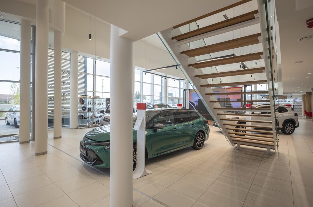 Wnętrze salonu samochodowego z zielonym kombi, dużymi oknami, białymi słupami i nowoczesnymi schodami