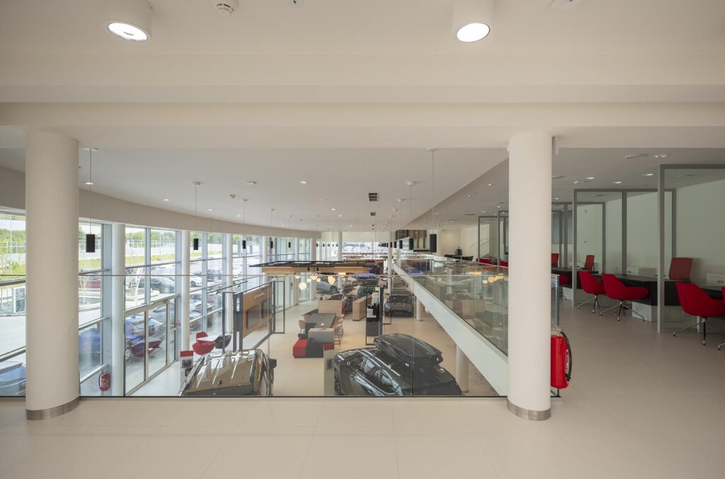 Przestronny, przeszklony salon samochodowy z wystawionymi autami i czerwonymi fotelami przy biurkach
