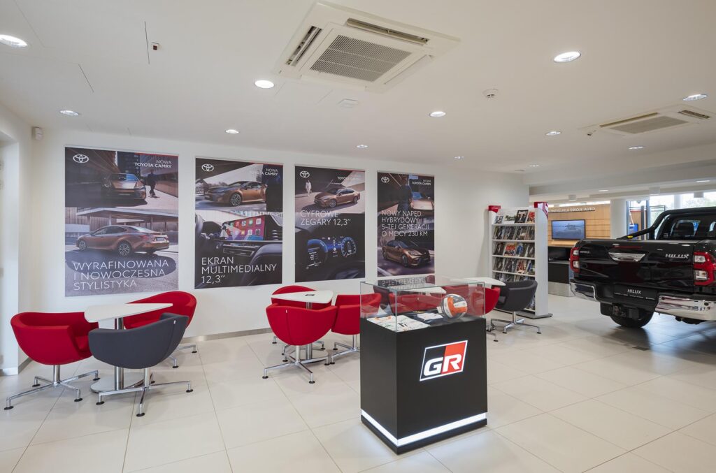 Wnętrze salonu samochodowego z czerwonymi fotelami, gablotą GR i wystawionym pickupem Hilux