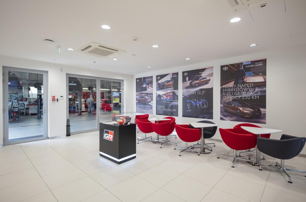Poczekalnia salonu samochodowego z czerwonymi fotelami, stolikami, stojakiem z logo GR i widokiem na halę serwisową przez przeszklone drzwi