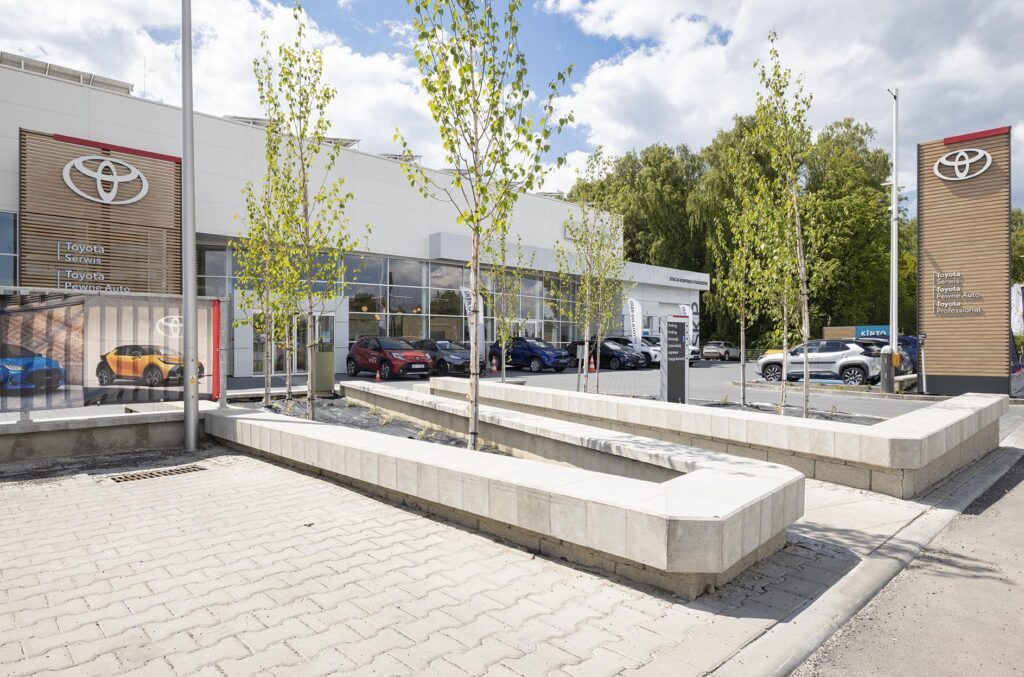 Salon Toyoty z dużym logo, zaparkowanymi samochodami i betonowymi donicami z młodymi drzewkami