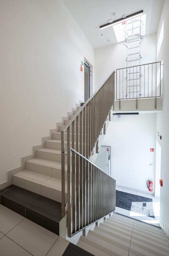 Jasna nowoczesna klatka schodowa z metalową balustradą, włazem dachowym z drabinką i gaśnicą