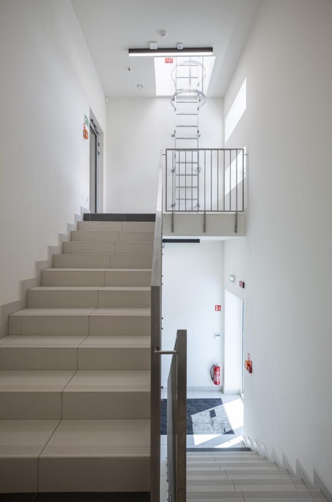 Nowoczesna jasna klatka schodowa z metalową balustradą, włazem dachowym i widoczną gaśnicą