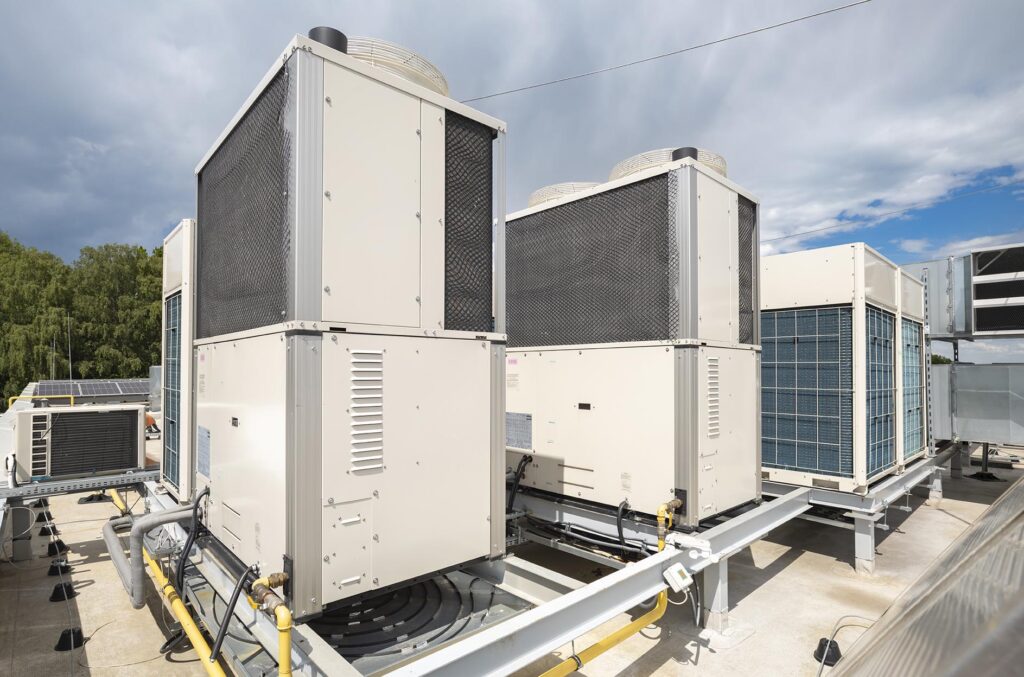 Agregaty klimatyzacyjne na dachu budynku na metalowych stelażach, rury i przewody, niebo i drzewa w tle